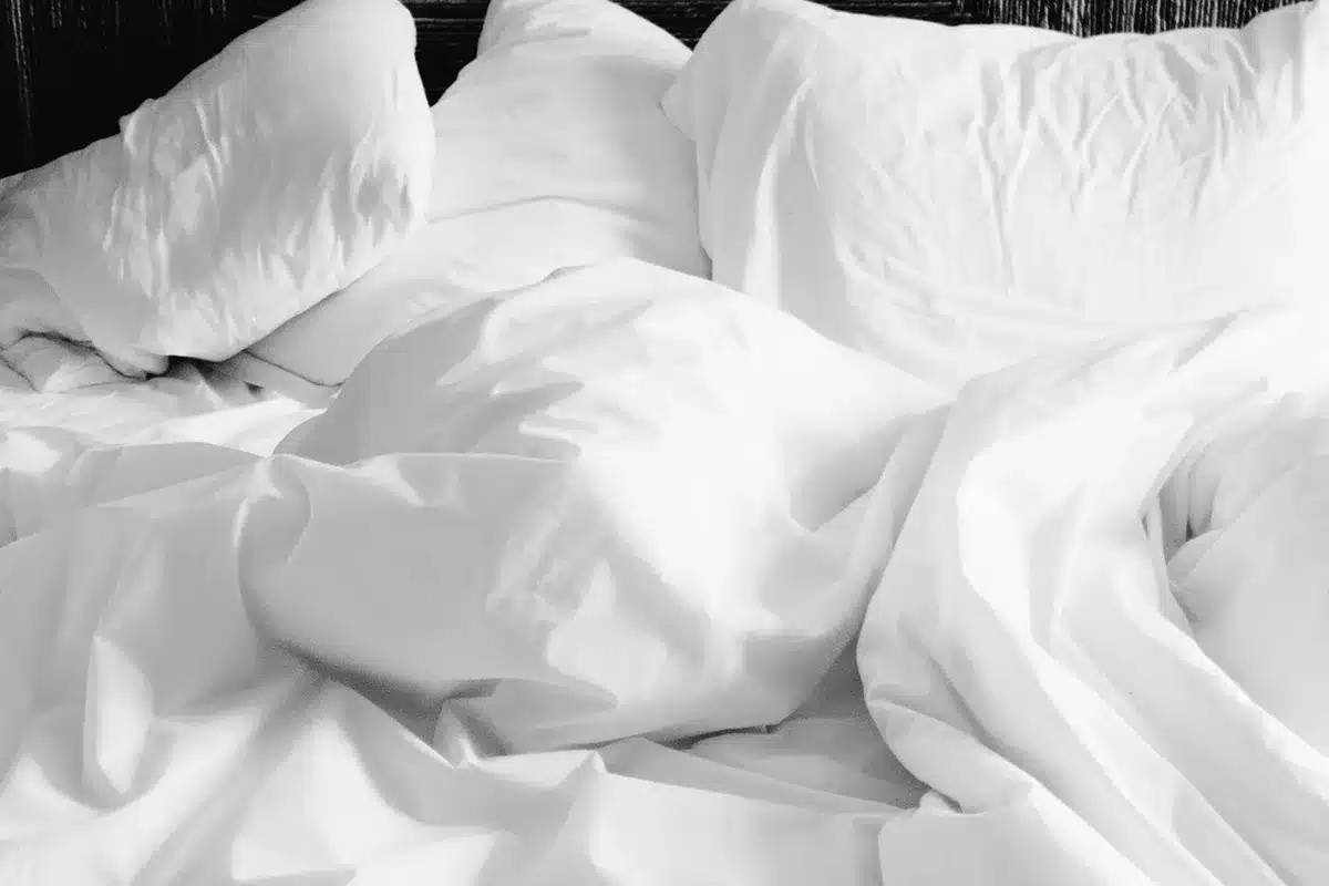 Chambre en plein jour avec draps légers en lin et percale, main touchant le tissu